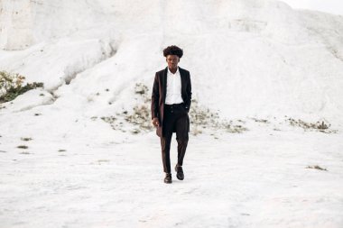 Siyah klasik takım elbiseli, modaya uygun siyah tenli Afro-Amerikalı adam, tebeşir dağlarının arka planında yürür ve poz verir, moda, erkek kıyafeti, poz verir