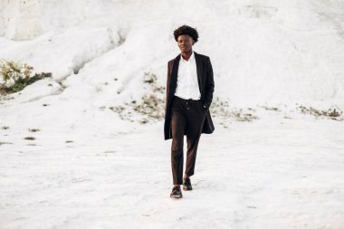 Siyah klasik takım elbiseli, modaya uygun siyah tenli Afro-Amerikalı adam, tebeşir dağlarının arka planında yürür ve poz verir, moda, erkek kıyafeti, poz verir