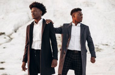 Klasik takım elbiseli ve hırkalı iki yakışıklı Afro-Amerikan erkeği, gün batımında güzel dağ manzarasının arka planında şık kıyafetler içinde poz veren iki genç adam.