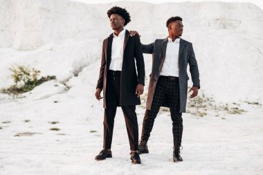 İki modaya uygun Afro-Amerikan erkeği, klasik siyah takım elbiseli ve polt giymiş, beyaz tebeşir dağlarının arka planına karşı birlikte poz veren adamlar, moda pozu veren konsept, erkek sonbahar kıyafetleri, tarz