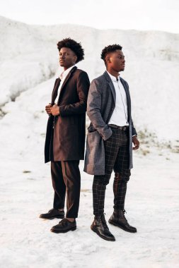 İki modaya uygun Afro-Amerikan erkeği, klasik siyah takım elbiseli ve polt giymiş, beyaz tebeşir dağlarının arka planına karşı birlikte poz veren adamlar, moda pozu veren konsept, erkek sonbahar kıyafetleri, tarz