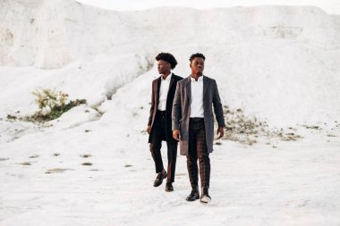 İki modaya uygun Afro-Amerikan erkeği, klasik siyah takım elbiseli ve polt giymiş, beyaz tebeşir dağlarının arka planına karşı birlikte poz veren adamlar, moda pozu veren konsept, erkek sonbahar kıyafetleri, tarz