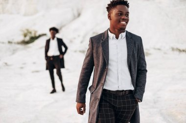 Sokakta poz veren iki manken, klasik kıyafetler, takım elbise, ceket, moda Afrikalı erkek modeller, kayaların arka planında.