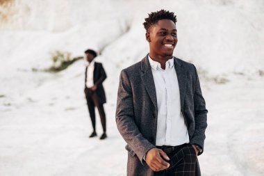Sokakta poz veren iki manken, klasik kıyafetler, takım elbise, ceket, moda Afrikalı erkek modeller, kayaların arka planında.