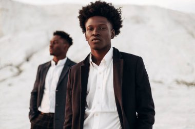 Sokakta poz veren iki manken, klasik kıyafetler, takım elbise, ceket, moda Afrikalı erkek modeller, kayaların arka planında.