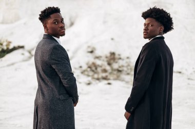 Sokakta poz veren iki manken, klasik kıyafetler, takım elbise, ceket, moda Afrikalı erkek modeller, kayaların arka planında.