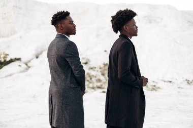 İki şık, siyah kıvırcık saçlı adam şık takım elbiseli, iki adam yan tarafa bakıyor, gün batımında kayalık tebeşir dağlarının zemininde poz veriyorlar.