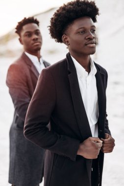 İki şık, siyah kıvırcık saçlı adam şık takım elbiseli, iki adam yan tarafa bakıyor, gün batımında kayalık tebeşir dağlarının zemininde poz veriyorlar.