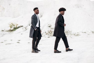 İki yakışıklı Afrikalı siyah adam, klasik giysiler içinde yürüyorlar ve poz veriyorlar, modaya uygun ceketlerle, gün batımında beyaz tebeşir dağlarının arka planında