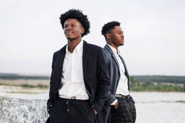 İki şık, siyah kıvırcık saçlı adam şık takım elbiseli, iki adam yan tarafa bakıyor, gün batımında kayalık tebeşir dağlarının zemininde poz veriyorlar.