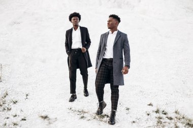 İki yakışıklı Afrikalı siyah adam, klasik giysiler içinde yürüyorlar ve poz veriyorlar, modaya uygun ceketlerle, gün batımında beyaz tebeşir dağlarının arka planında