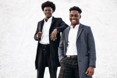 Sokakta poz veren iki manken, klasik kıyafetler, takım elbise, ceket, moda Afrikalı erkek modeller, kayaların arka planında.