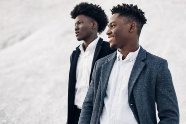 Sokakta poz veren iki manken, klasik kıyafetler, takım elbise, ceket, moda Afrikalı erkek modeller, kayaların arka planında.