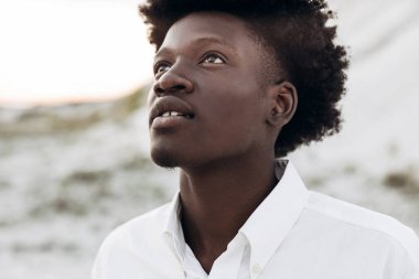 Havalı, kıvırcık, yakışıklı, dışarıda dikilen siyahi bir adamın portresi, modaya uygun Afro-Amerikan bir adam, kayaların arkasına bakıyor ve poz veriyor.