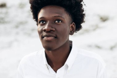 Havalı, kıvırcık, yakışıklı, dışarıda dikilen siyahi bir adamın portresi, modaya uygun Afro-Amerikan bir adam, kayaların arkasına bakıyor ve poz veriyor.