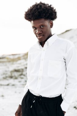 Havalı, kıvırcık, yakışıklı, dışarıda dikilen siyahi bir adamın portresi, modaya uygun Afro-Amerikan bir adam, kayaların arkasına bakıyor ve poz veriyor.