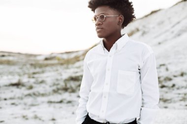 Beyaz bir gömlek ve pantolon giyen kıvırcık saçlı yakışıklı Afro-Amerikalı bir adam, kayalık dağların manzarasına karşı sokakta poz veriyor, moda, moda...