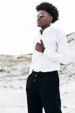 Beyaz bir gömlek ve pantolon giyen kıvırcık saçlı yakışıklı Afro-Amerikalı bir adam, kayalık dağların manzarasına karşı sokakta poz veriyor, moda, moda...