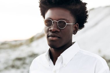 Moda fotoğrafı, yakışıklı kıvırcık saçlı, güneş gözlüklü, zarif bir gömlek giyen, Eski Şehir 'deki kayalık dağların yakınında poz veren siyah Afrikalı bir model, erkek tarzı, güzellik.