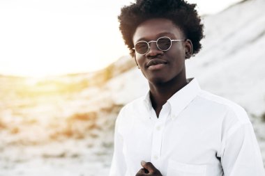 Moda fotoğrafı, yakışıklı kıvırcık saçlı, güneş gözlüklü, zarif bir gömlek giyen, Eski Şehir 'deki kayalık dağların yakınında poz veren siyah Afrikalı bir model, erkek tarzı, güzellik.