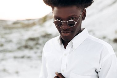 Moda fotoğrafı, yakışıklı kıvırcık saçlı, güneş gözlüklü, zarif bir gömlek giyen, Eski Şehir 'deki kayalık dağların yakınında poz veren siyah Afrikalı bir model, erkek tarzı, güzellik.
