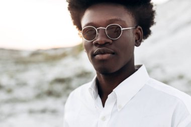 Moda fotoğrafı, yakışıklı kıvırcık saçlı, güneş gözlüklü, zarif bir gömlek giyen, Eski Şehir 'deki kayalık dağların yakınında poz veren siyah Afrikalı bir model, erkek tarzı, güzellik.