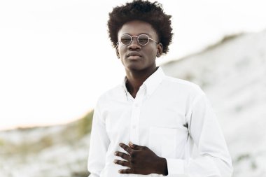 Erkek portresi, yakışıklı kıvırcık saçlı, beyaz gömlek ve güneş gözlüğü takan, açık havada gülümseyen Amerikalı.