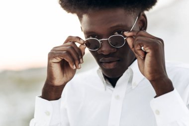 Erkek portresi, yakışıklı kıvırcık saçlı, beyaz gömlek ve güneş gözlüğü takan, açık havada gülümseyen Amerikalı.