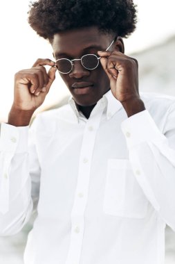 Erkek portresi, yakışıklı kıvırcık saçlı, beyaz gömlek ve güneş gözlüğü takan, açık havada gülümseyen Amerikalı.