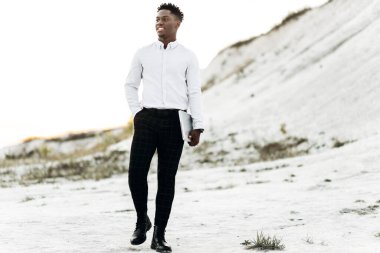 Yakışıklı, zarif, beyaz gömlekli siyah adam, beyaz tebeşir kayaları ve dağların arka planına karşı dizüstü bilgisayarını taşıyor, moda, iş, tarz kavramı