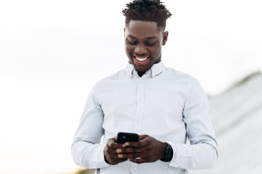 Beyaz gömlekli, neşeli, kıvırcık Afrikalı adam, cep telefonu kullanıyor, sokakta, iş konsepti, iş yeri, aletler.