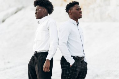 Beyaz gömlekli ve güneş gözlüklü iki yakışıklı adam, sırt sırta verip, taşlı beyaz dağların arkasında poz veriyorlar, şık Afrikalı erkekler, şık pozlar, erkek kıyafetleri..