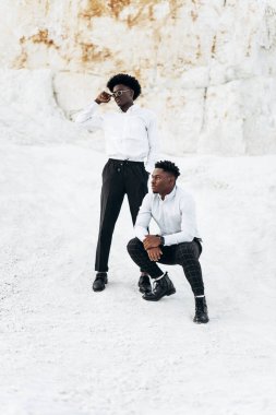 İki zarif siyah Afrikalı, beyaz gömlekli, tam gelişim içinde, dağların ve kayaların yanında açık havada poz veriyorlar, arkasında güzel bir manzara olan moda, moda pozu veriyorlar.,