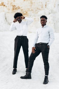İki zarif siyah Afrikalı, beyaz gömlekli, tam gelişim içinde, dağların ve kayaların yanında açık havada poz veriyorlar, arkasında güzel bir manzara olan moda, moda pozu veriyorlar.,