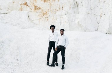 İki şık, yakışıklı Afro-Amerikan erkek. Siyah gözlükler ve beyaz zarif gömlekler içinde, kayalık bir bölgede dağların arka planına karşı poz veren modeller..