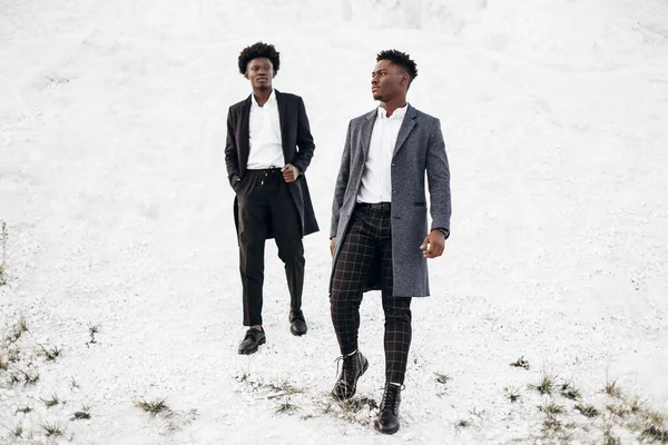 İki yakışıklı Afrikalı siyah adam, klasik giysiler içinde yürüyorlar ve poz veriyorlar, modaya uygun ceketlerle, gün batımında beyaz tebeşir dağlarının arka planında