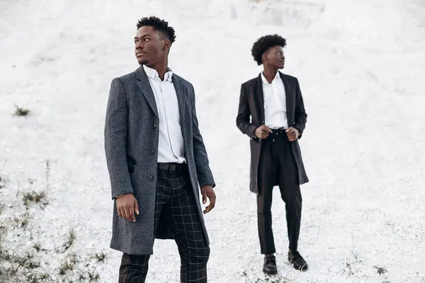 Havalı Afro-Amerikan kıvırcık saçlı, şık, benzer takım elbiseli adamlar, gün batımında kayalık bir bölgede moda pozu veriyorlar.