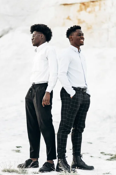Two handsome fashionable American men, in white shirts and sunglasses, posing back to back, against the backdrop of stony white mountains, stylish African men, fashionable posing, men\'s clothing.