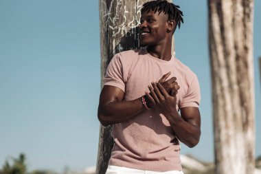 Çöl bölgesinde tahta sütunların yanında dururken gülümseyen yakışıklı siyah adamın portresi, moda anlayışı, insanlar, Afrika doğası, kum tepeleri.