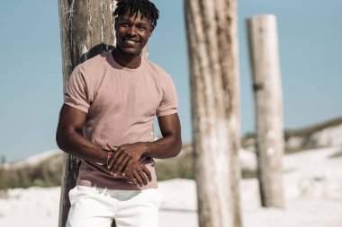 Çöl bölgesinde tahta sütunların yanında dururken gülümseyen yakışıklı siyah adamın portresi, moda anlayışı, insanlar, Afrika doğası, kum tepeleri.