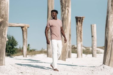 Neşeli gülümseyen stil Afrikalı, pembe tişörtlü ve beyaz pantolonlu adam, Afro-Amerikan model adam, kuru çölde yürüyor, moda kavramı, insanlar, Vahşi Batı tarzı.