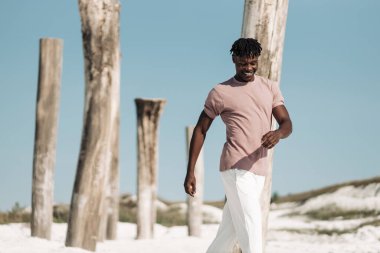 Neşeli gülümseyen stil Afrikalı, pembe tişörtlü ve beyaz pantolonlu adam, Afro-Amerikan model adam, kuru çölde yürüyor, moda kavramı, insanlar, Vahşi Batı tarzı.