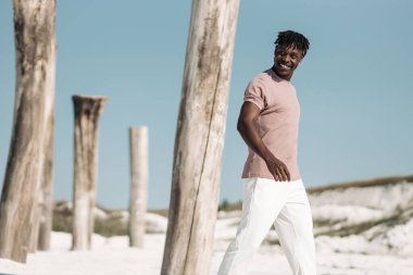 Yakışıklı Afrikalı bir adam, gülümseyen ve kameraya bakan, kumlu çölde mavi gökyüzü arka planında yürüyen, tahta sütunları olan, manken konsepti olan,