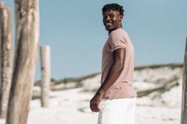 Yakışıklı Afrikalı bir adam, gülümseyen ve kameraya bakan, kumlu çölde mavi gökyüzü arka planında yürüyen, tahta sütunları olan, manken konsepti olan,