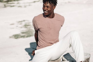 Moda ve şık kıyafetlerle poz veren şık, kıvırcık siyah Afrikalı Amerikalı adam kumların üzerinde oturuyor, moda kavramı, insanlar, tarz.