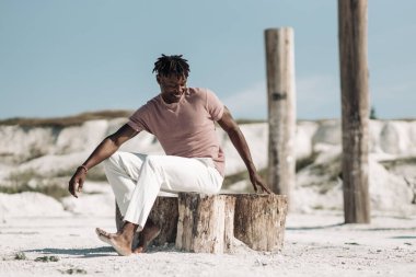 Tarz sahibi yakışıklı bir adamın açık bir portresi, şık kıyafetler içinde, kumda poz veren siyah bir adam, erkek moda konsepti.