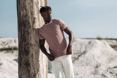 Tarz sahibi yakışıklı bir adamın açık bir portresi, şık kıyafetler içinde, kumda poz veren siyah bir adam, erkek moda konsepti.