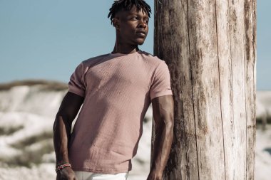 Tarz sahibi yakışıklı bir adamın açık bir portresi, şık kıyafetler içinde, kumda poz veren siyah bir adam, erkek moda konsepti.