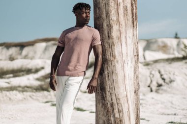 Tarz sahibi yakışıklı bir adamın açık bir portresi, şık kıyafetler içinde, kumda poz veren siyah bir adam, erkek moda konsepti.