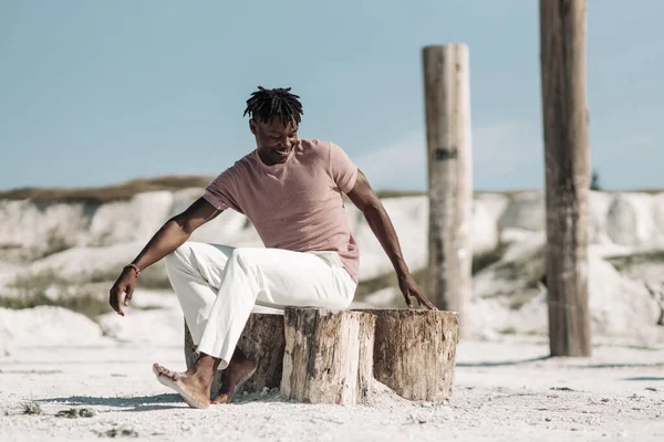 Tarz sahibi yakışıklı bir adamın açık bir portresi, şık kıyafetler içinde, kumda poz veren siyah bir adam, erkek moda konsepti.